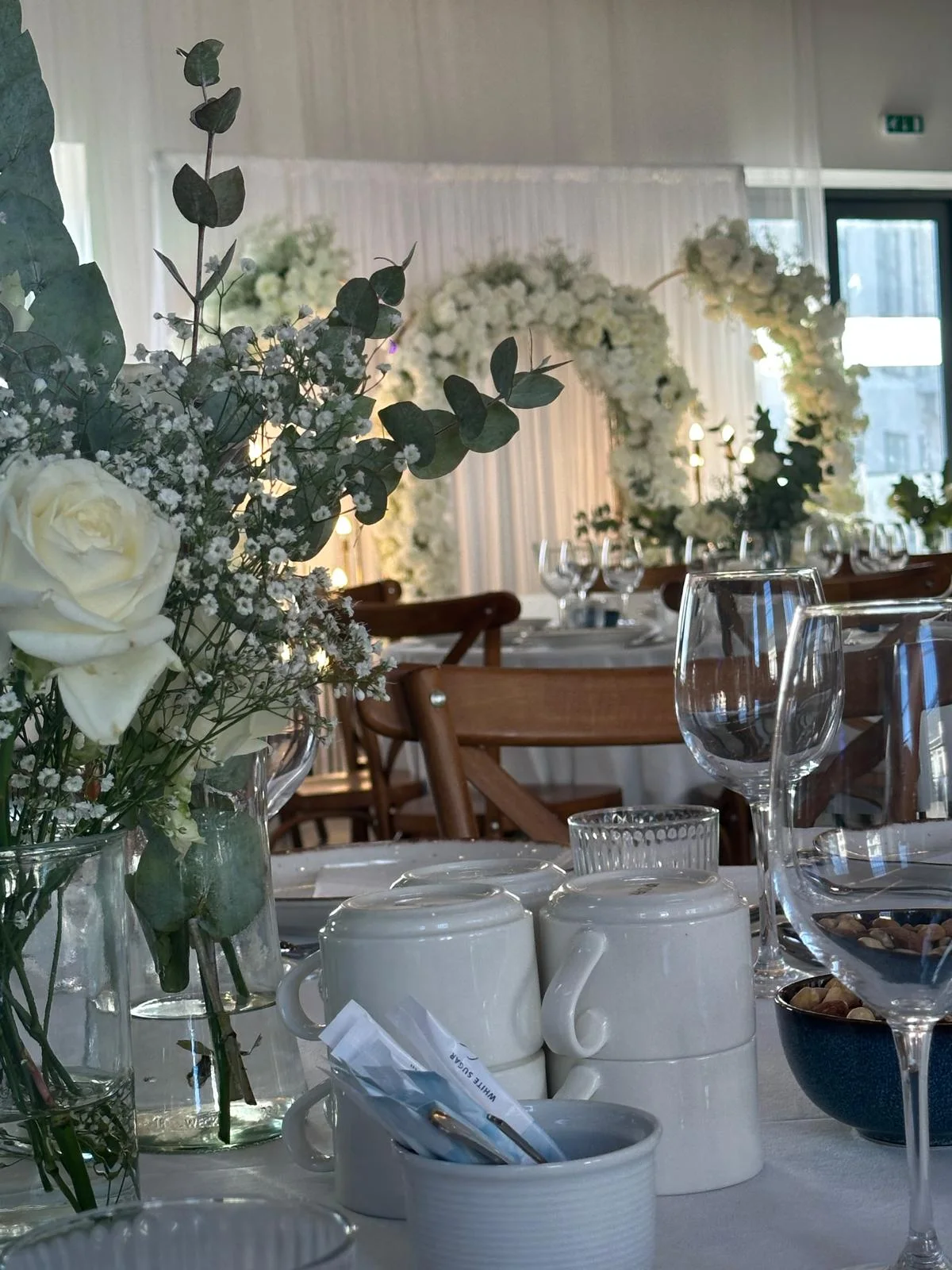 Hvide blomster og grønne grene på et festbord med vinglas og hvide duge